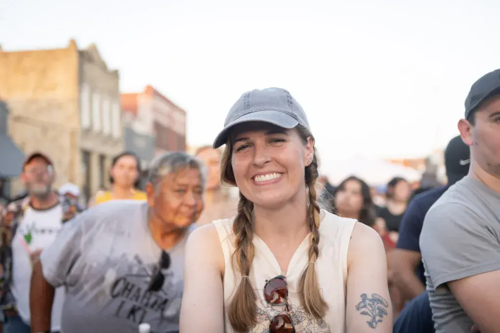 A smiling woman with braided hair and a blue cap stands among festival attendees with historic buildings in the background.