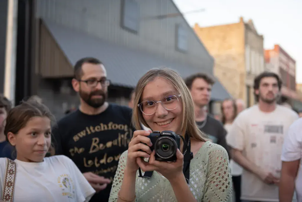 A young person wearing glasses holds up a camera while smiling at a crowded outdoor festival venue.