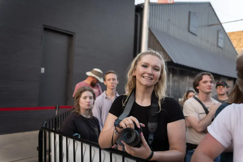 A smiling person holds a camera while standing among festival attendees near venue buildings.