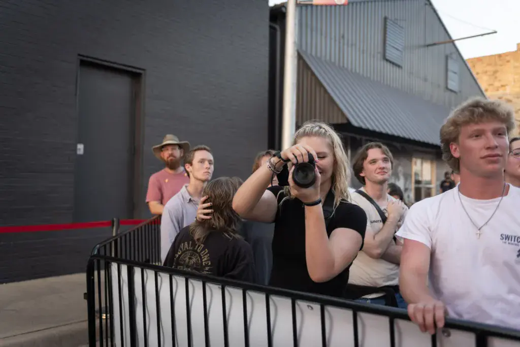 Festival attendees stand behind a barrier, with a woman in the center taking photos with a camera.