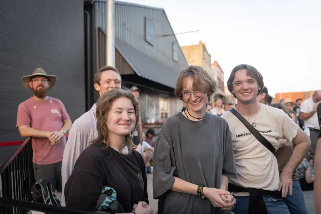 A group of four smiling festival attendees stand together outdoors near a metal structure at the venue.