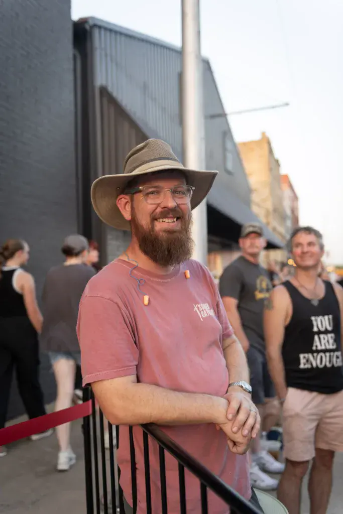 A man wearing a cowboy hat and pink t-shirt smiles while leaning against a barrier at an outdoor festival venue.