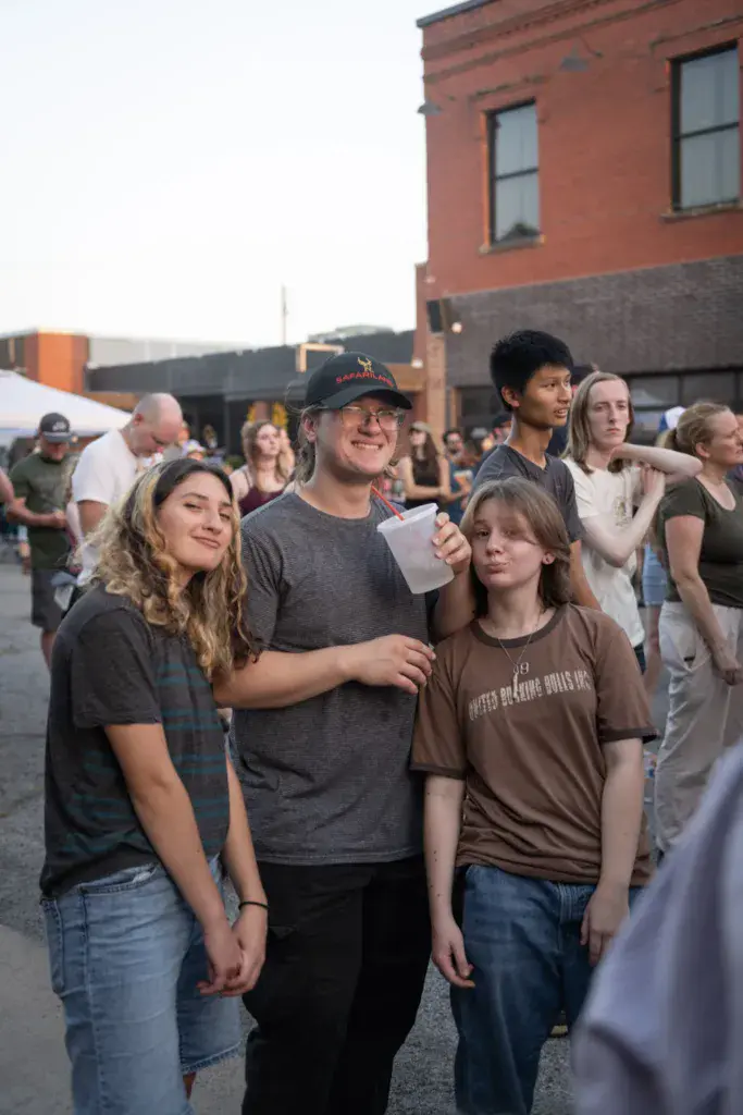 Festival attendees stand together in an outdoor venue area near a red brick building, smiling and looking upward.