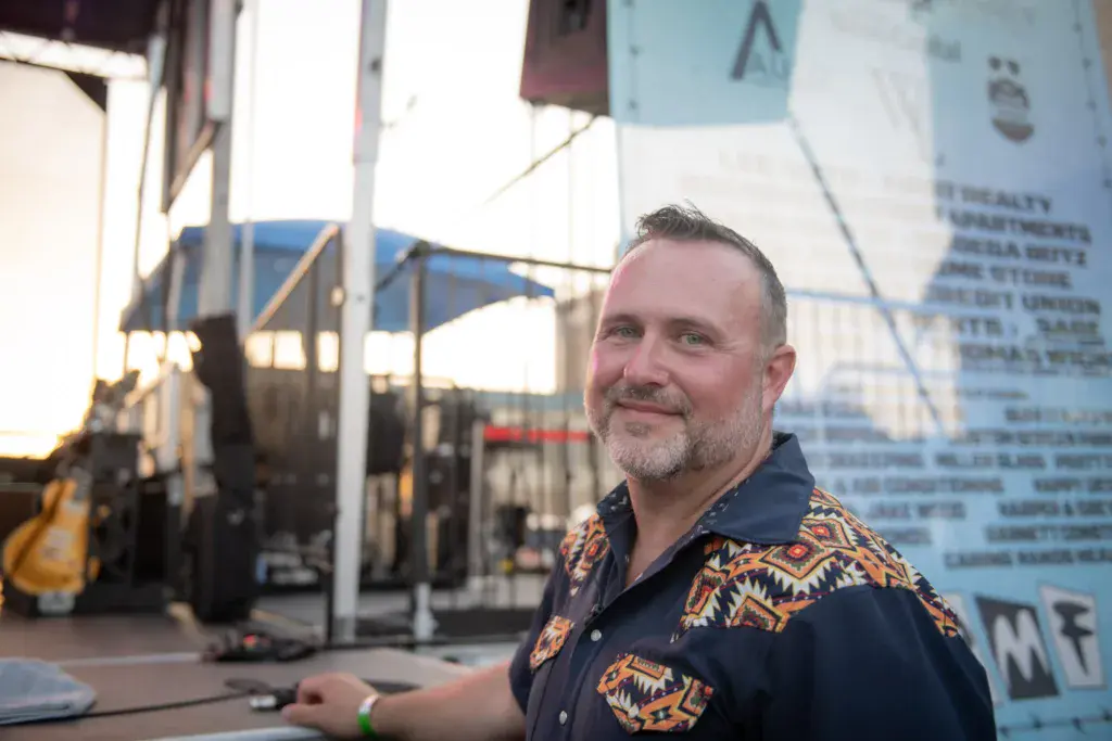 A person in a patterned black shirt stands near the stage area at an outdoor music festival venue.