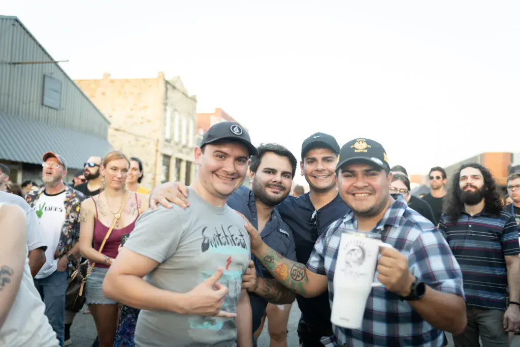 A group of smiling men pose together at an outdoor festival with historic buildings in the background.