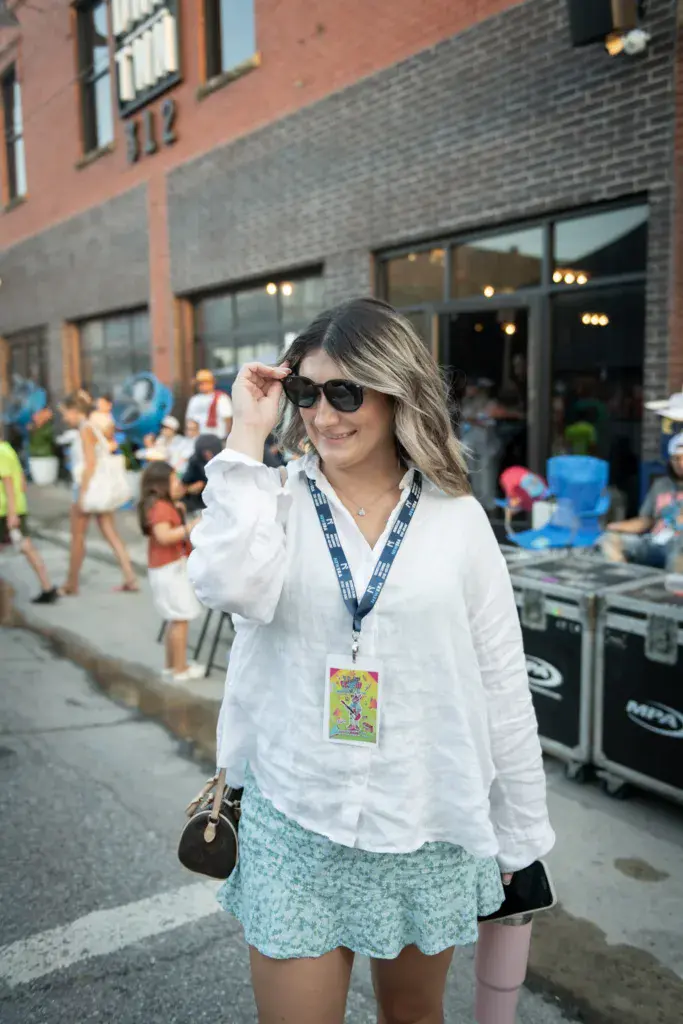 Festival attendee wearing lanyard badge and sunglasses smiles at camera with brick venue building and crowd in background.