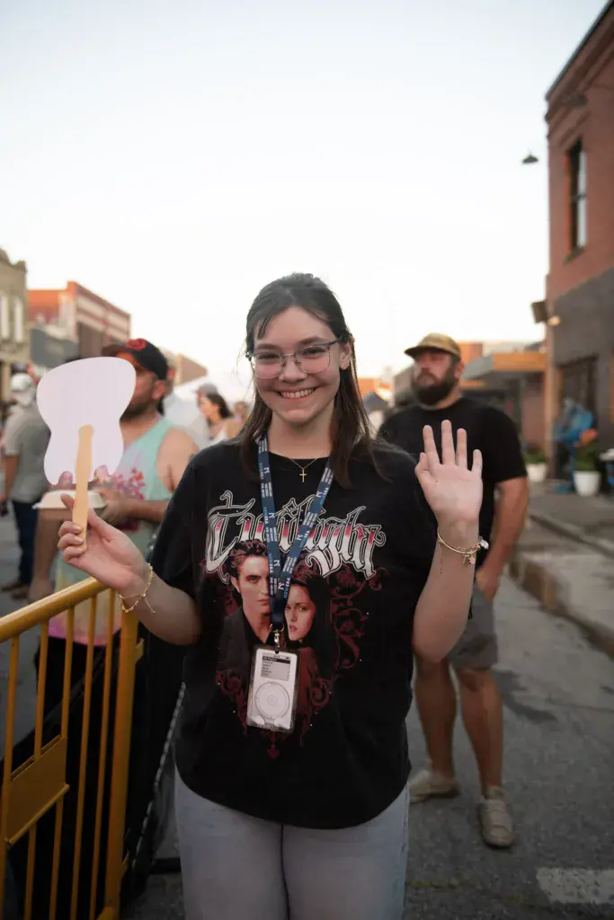 A smiling attendee wearing a festival badge and vintage band shirt holds a foam guitar cutout while waving at the Dancing Rabbit Music Festival.