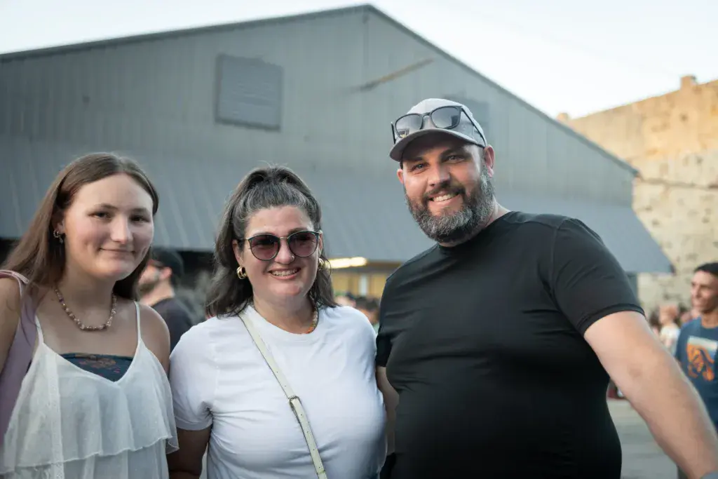 Three smiling festival attendees pose together in front of a large metal building at the venue.