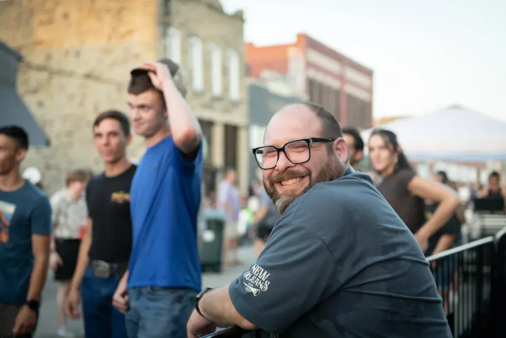 A smiling man wearing glasses and a gray shirt turns toward the camera at an outdoor festival with other attendees and historic buildings in the background.