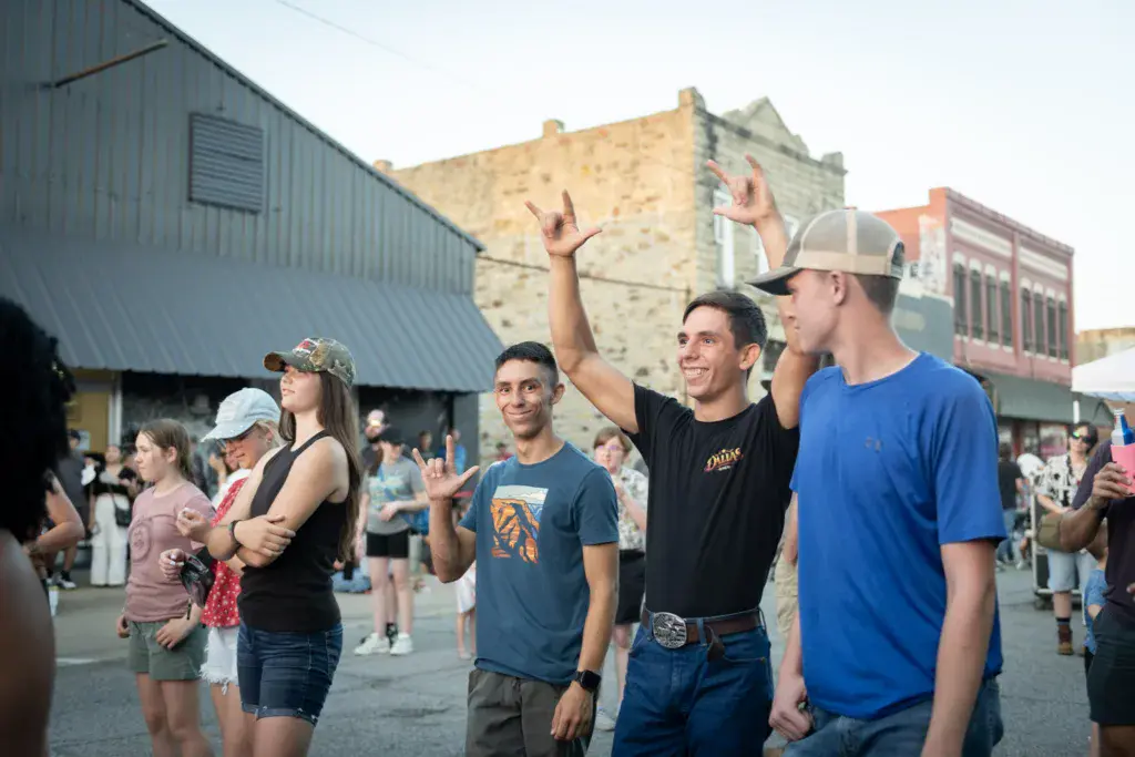 A group of smiling attendees celebrates at the Dancing Rabbit Music Festival with brick buildings and crowds in the background.