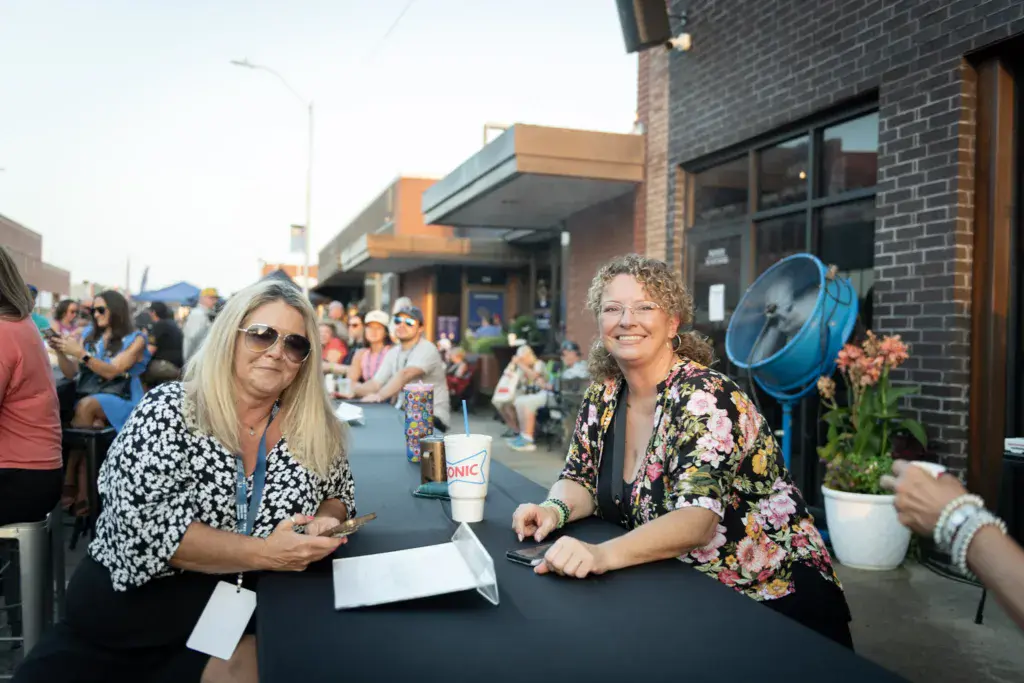 Two women sit at a table at an outdoor festival venue with crowds and brick buildings visible behind them.
