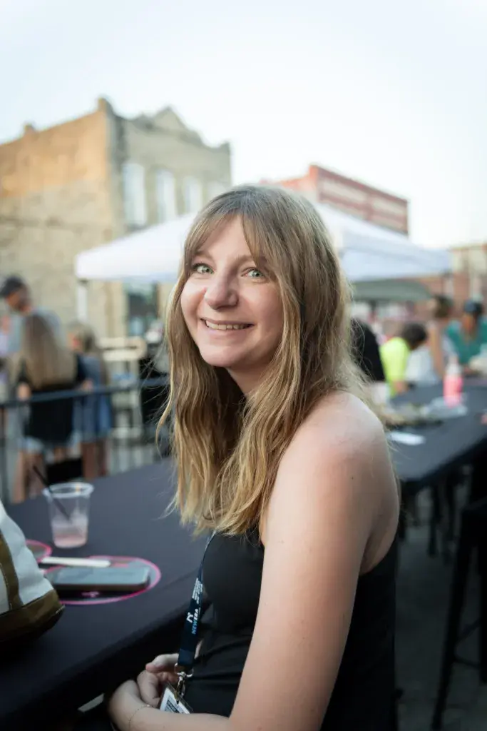 A smiling woman at an outdoor festival event with crowds and historic buildings in the background.