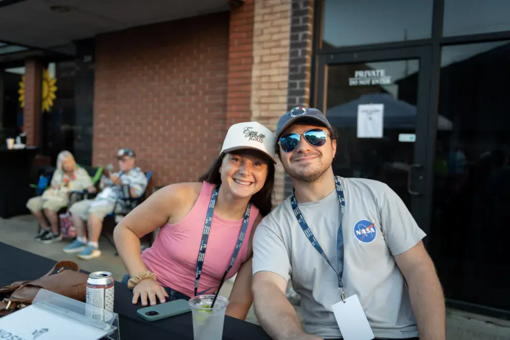 Two festival attendees wearing lanyards smile together at an outdoor venue with brick buildings and other guests visible behind them.
