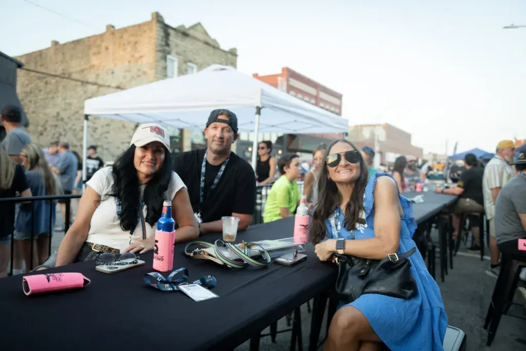 Festival attendees sit at a table under a white tent at an outdoor urban venue.