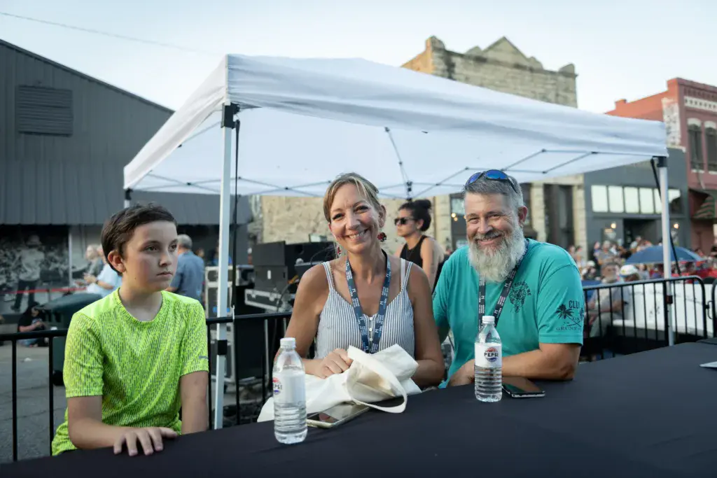 Three people sit at a table under a white tent at an outdoor music festival venue.