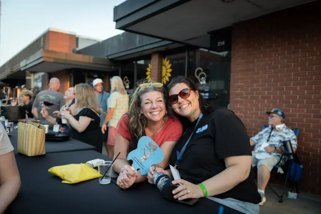 Two smiling festival-goers pose together at an outdoor venue with festival buildings and crowds in the background.