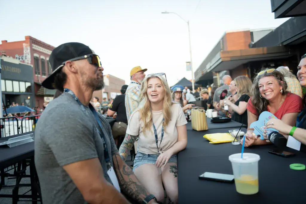 Festival attendees enjoy beverages and conversation at an outdoor table with urban buildings in the background.