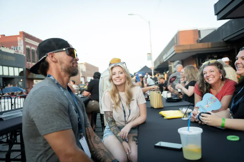 Festival attendees socialize at an outdoor table with drinks and snacks, with brick buildings visible in the background.