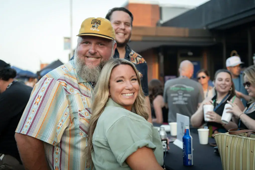 Smiling festival attendees pose together at an outdoor music event with crowds and venue structures visible behind them.