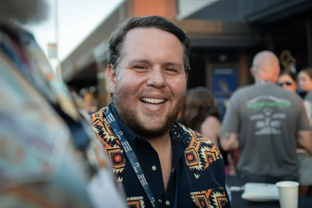 A man in a colorful patterned shirt smiles at the Dancing Rabbit Music Festival surrounded by festival attendees.