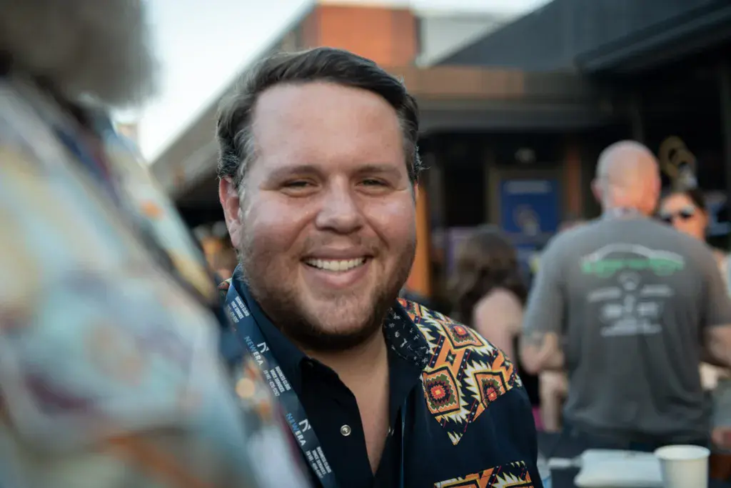 A person smiling at the camera wearing a festival lanyard and patterned black shirt at an outdoor event.