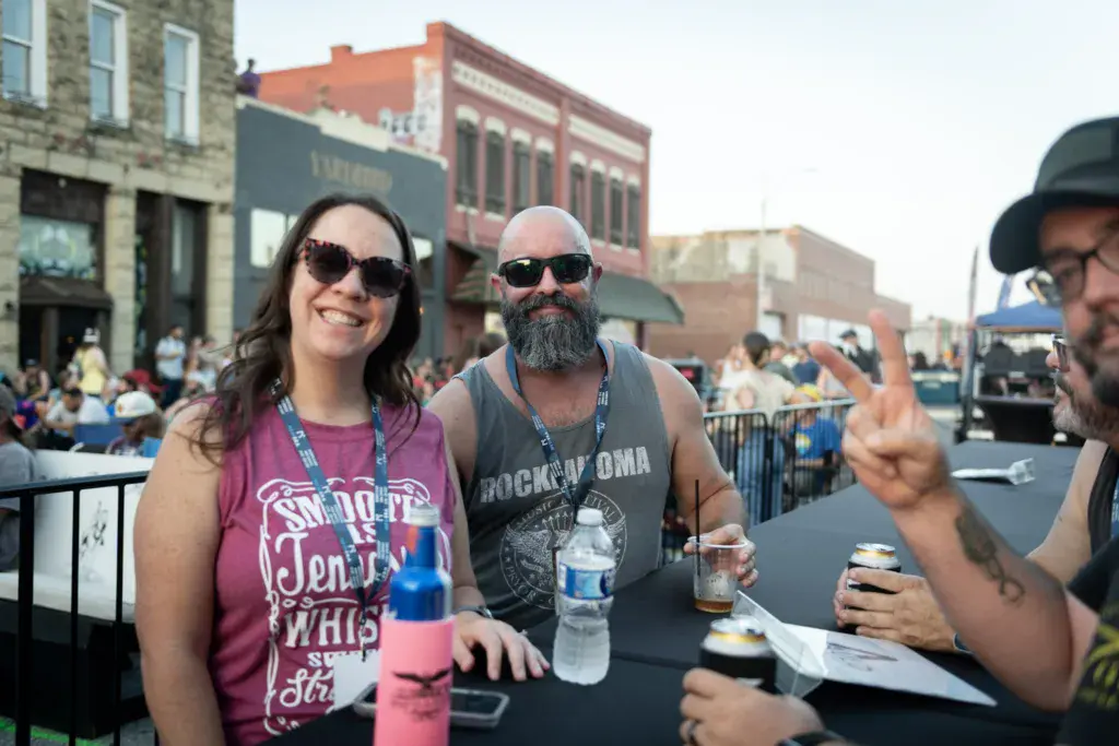 Festival attendees enjoy drinks and socialize at an outdoor music event with historic brick buildings in the background.