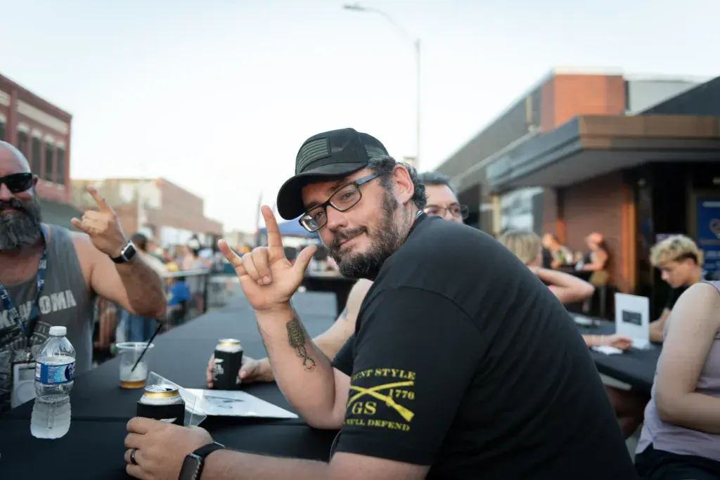 A person wearing glasses and a black cap makes a peace sign while sitting at a table with drinks during an outdoor festival.