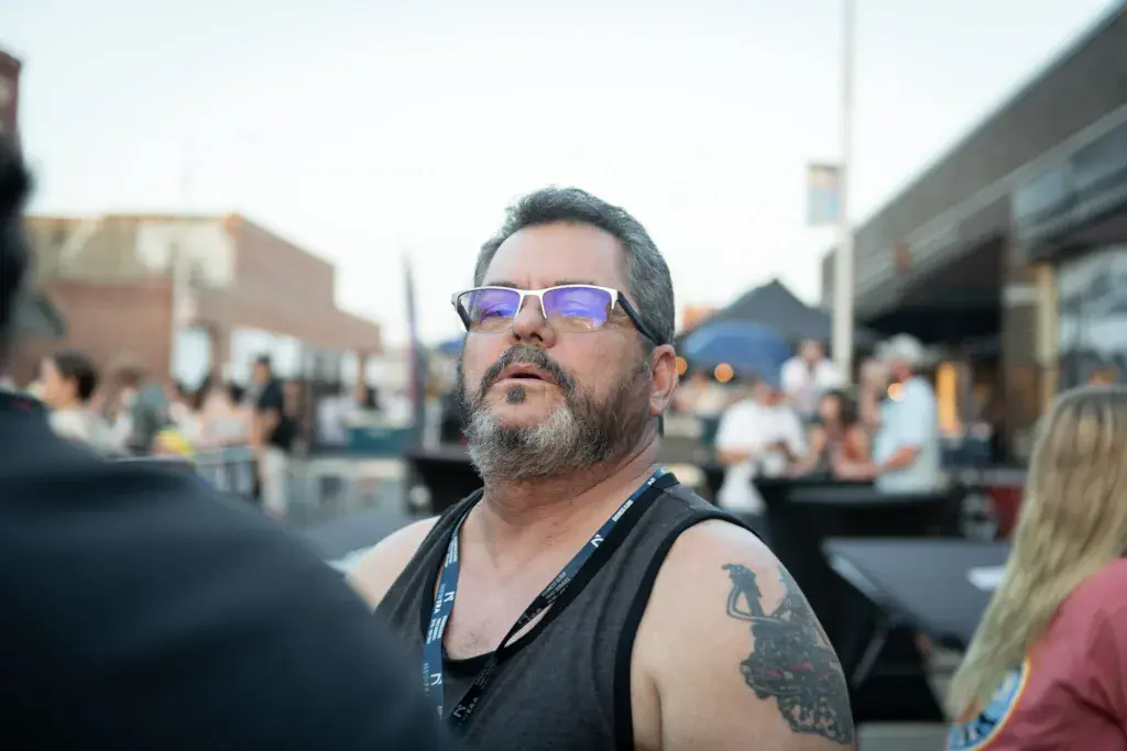 A man wearing sunglasses and a lanyard stands in a festival crowd with outdoor venue structures visible in the background.