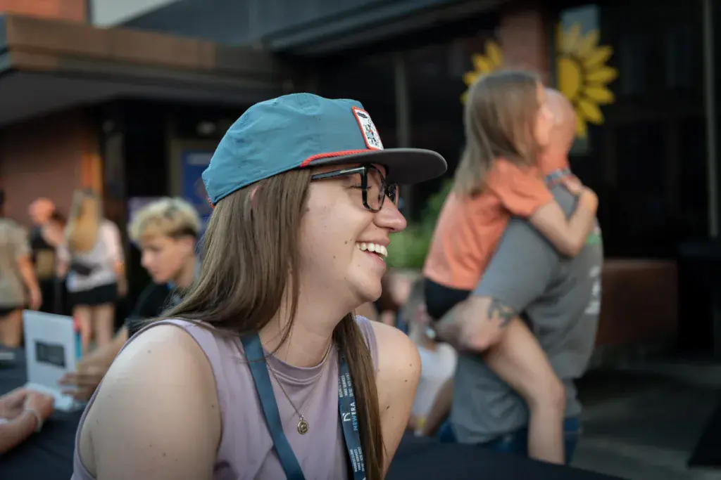 A smiling person wearing a blue and red cap and festival lanyard stands in a crowd at an outdoor venue.