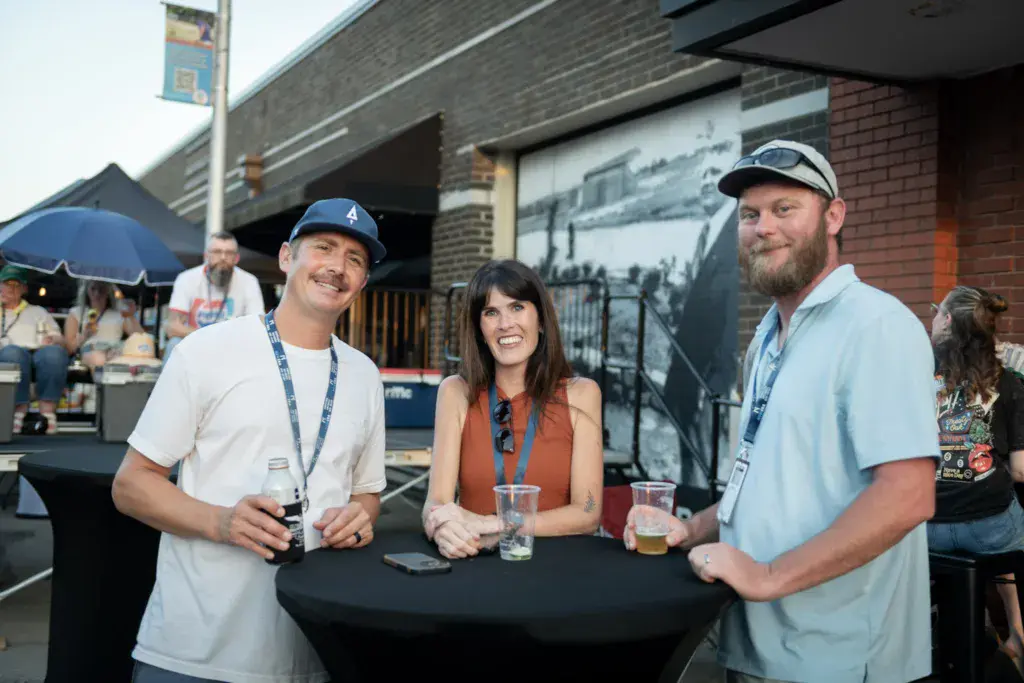 Three people wearing festival lanyards smile while holding drinks at an outdoor venue with brick buildings.