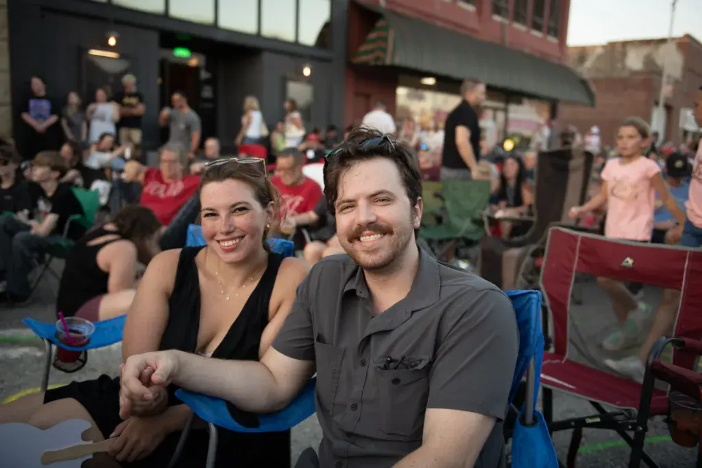 Two smiling attendees sit together in blue camp chairs at an outdoor festival venue with crowds visible behind them.