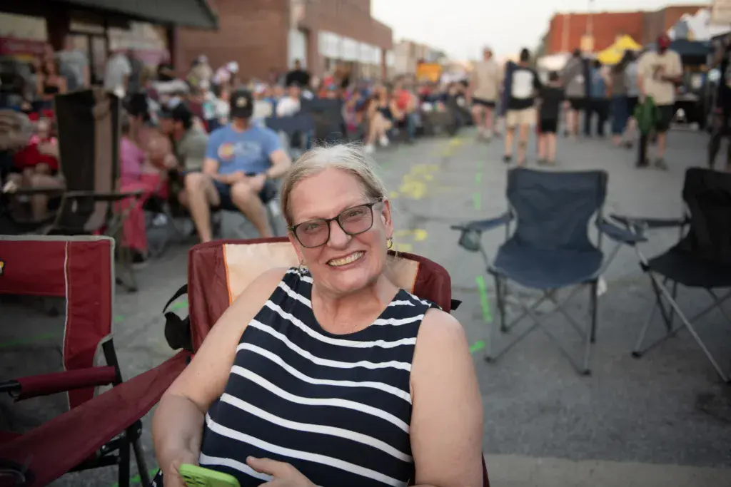 A smiling woman wearing glasses and a striped shirt sits in a red camping chair at an outdoor festival surrounded by crowds.