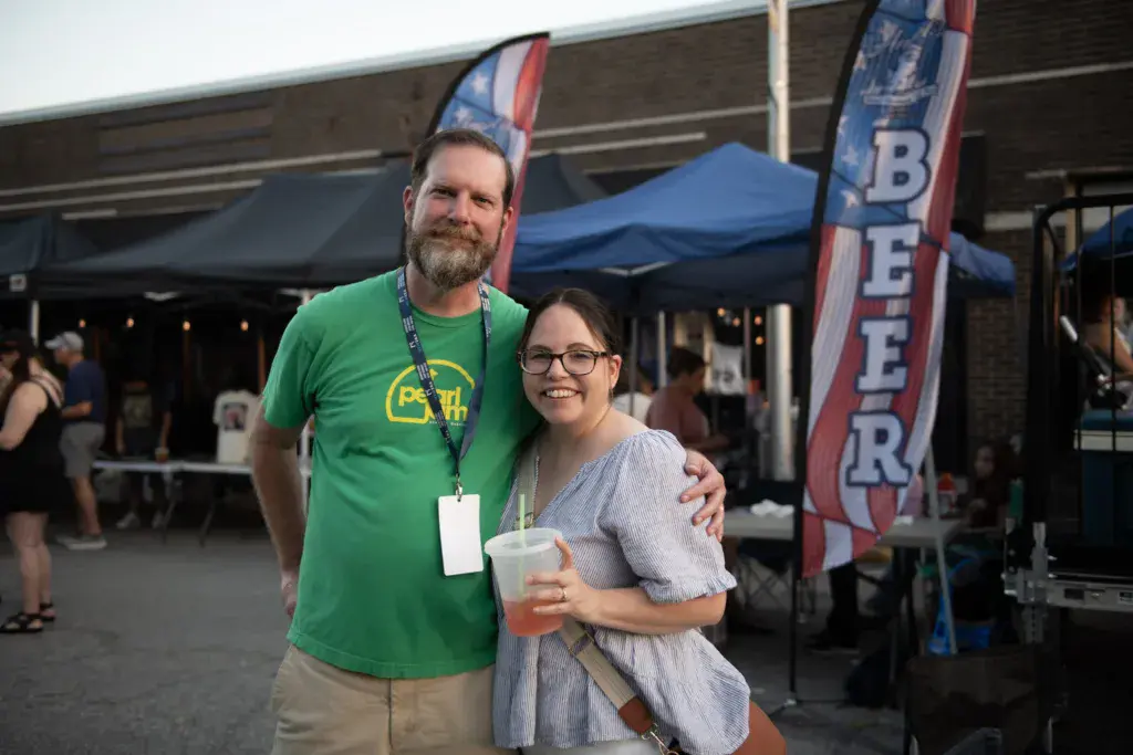 Two people pose together at an outdoor festival venue with tents and signage visible behind them.