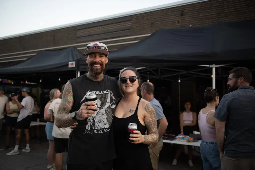 Two festival attendees smile while holding drinks under a tent canopy at an outdoor music event.