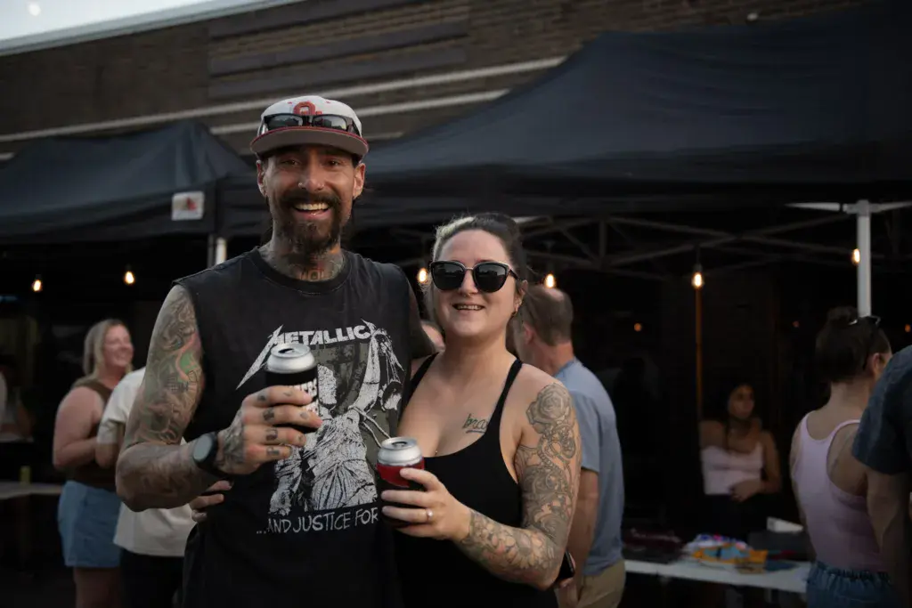 Two festival attendees smile while holding drinks under a covered outdoor venue at dusk.