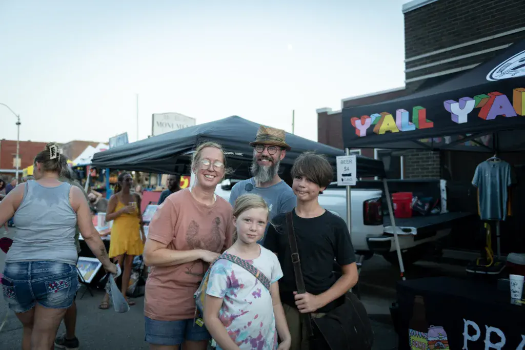 A family of four poses together at an outdoor festival with vendor tents and colorful signage in the background.