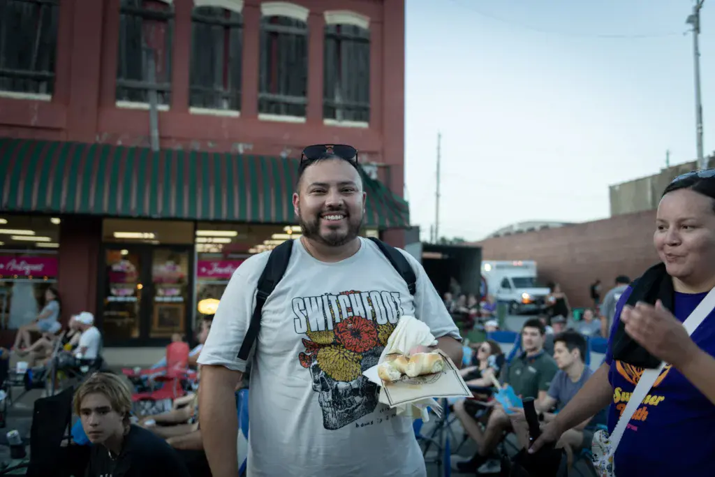 A smiling festival attendee holds food while standing in a crowded outdoor venue with brick buildings.
