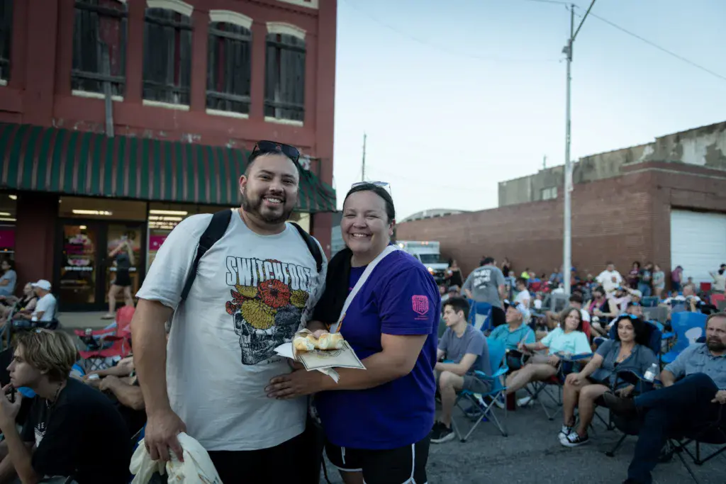 Two smiling festival attendees holding food stand in front of a seated crowd at an outdoor music festival.