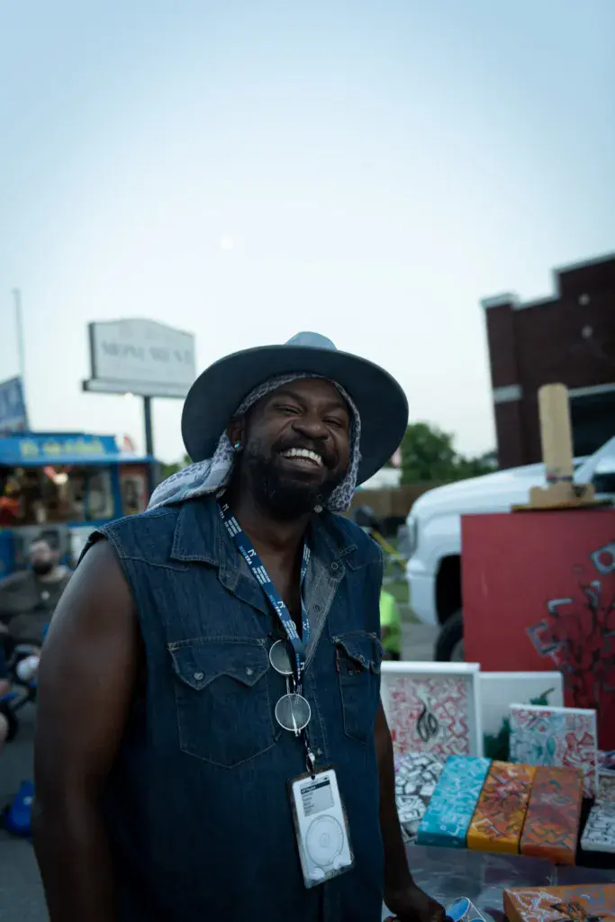 A smiling person wearing a black cowboy hat and denim vest stands at a vendor booth displaying colorful merchandise.