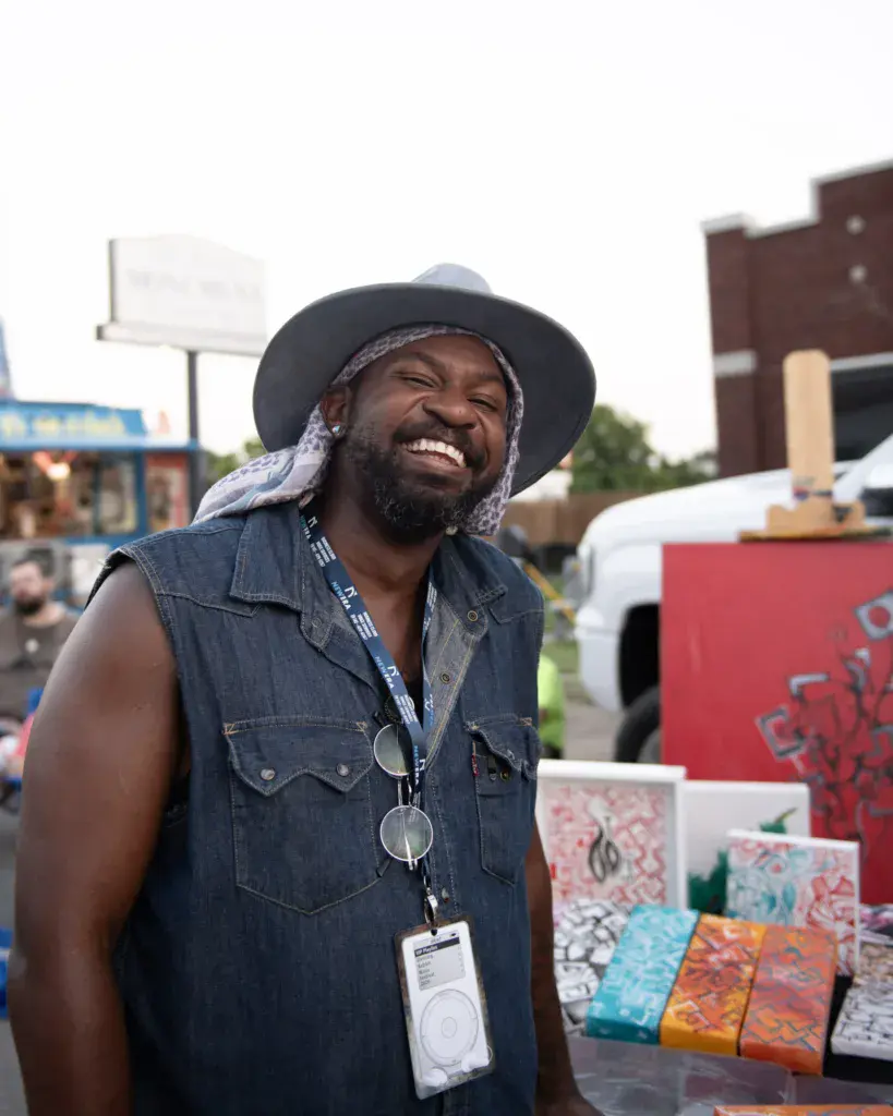 A smiling person wearing a black cowboy hat and denim vest with a festival lanyard stands at an outdoor vendor booth.