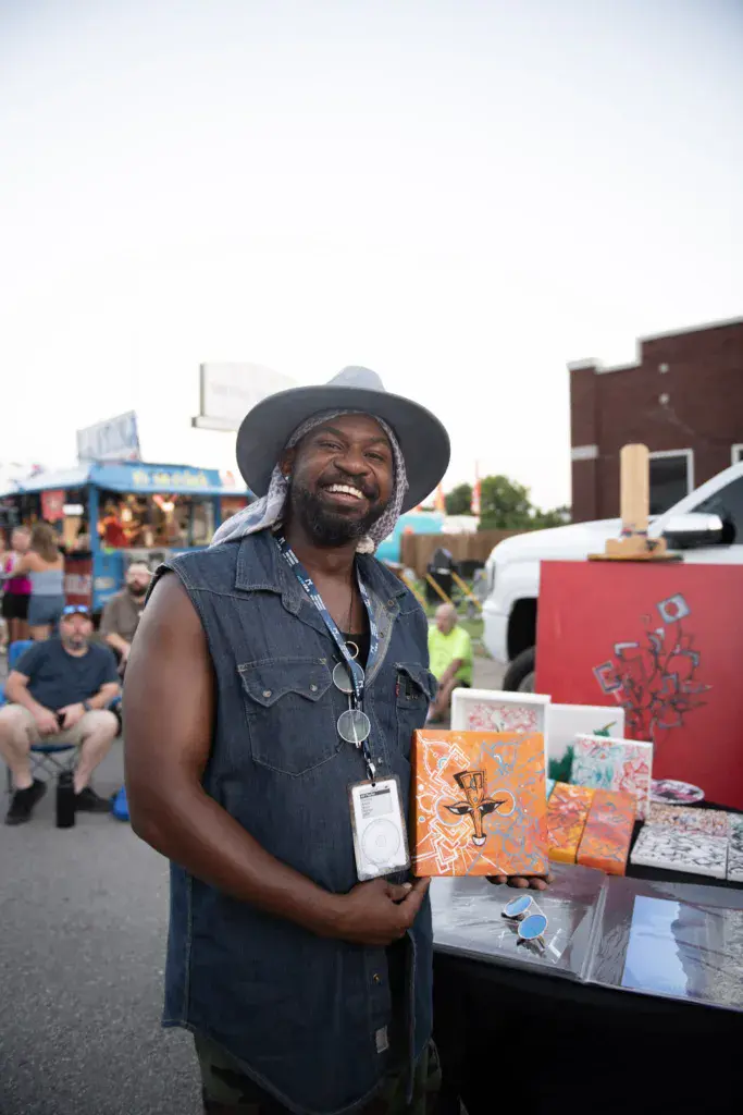 A smiling vendor at the Dancing Rabbit Music Festival displays artwork and merchandise at their booth.