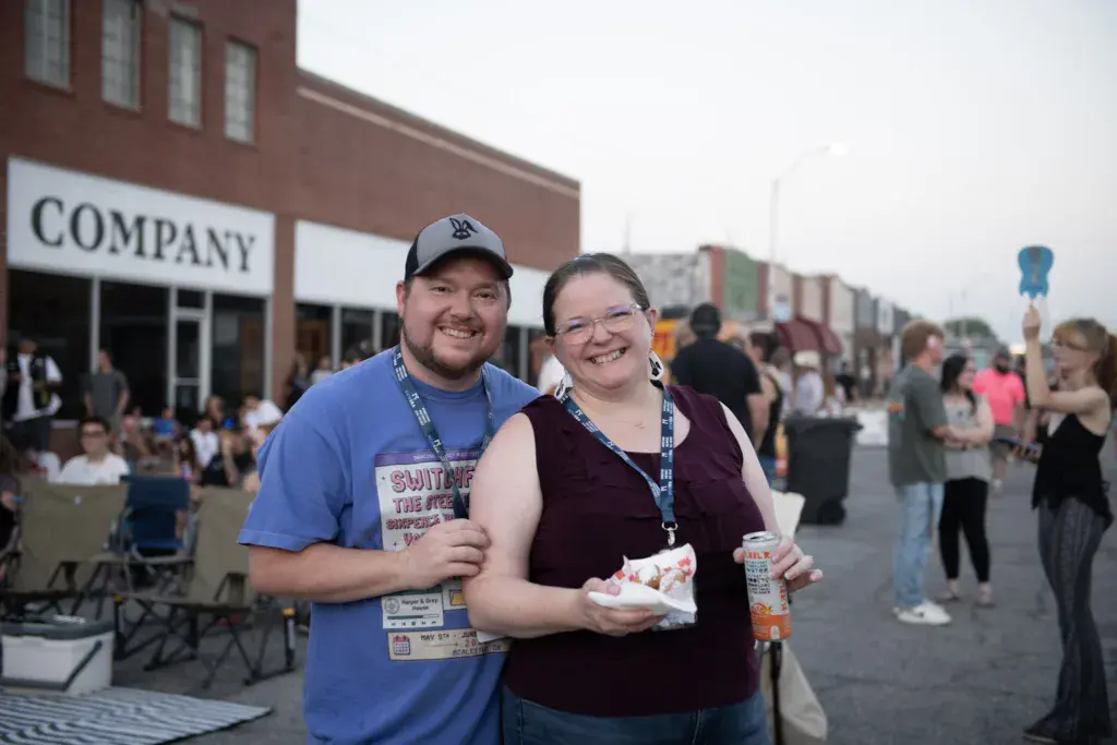 Two smiling festival attendees with lanyards hold food and drinks in front of brick buildings and festival crowds.