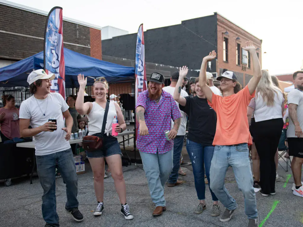 A group of festival attendees dancing and celebrating together in an outdoor venue area with a blue tent and brick buildings in the background.