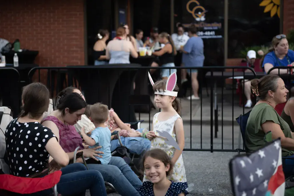 Families with children enjoy outdoor seating at a music festival venue with a stage visible in the background.