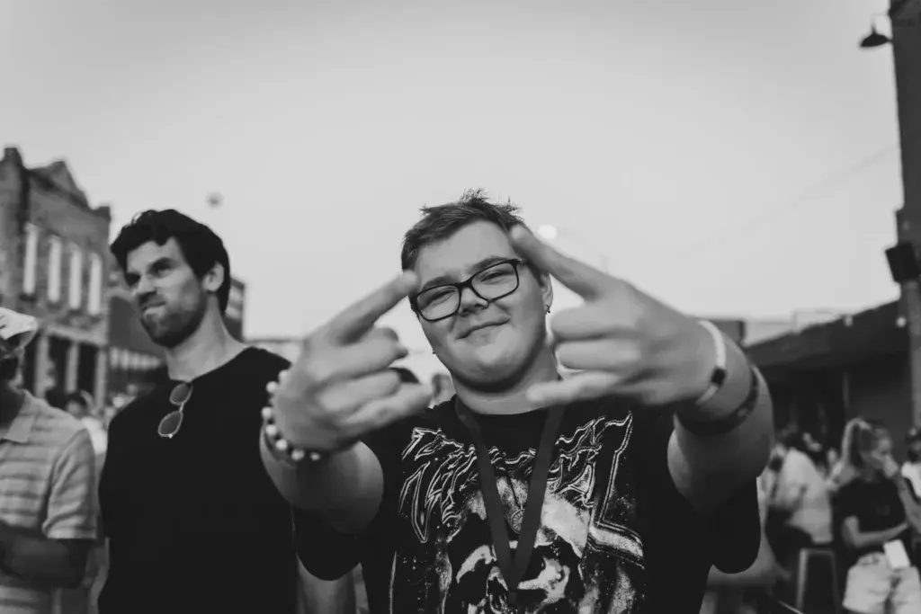 A person in glasses making a playful pointing gesture at the camera while standing in a crowd at an outdoor festival.