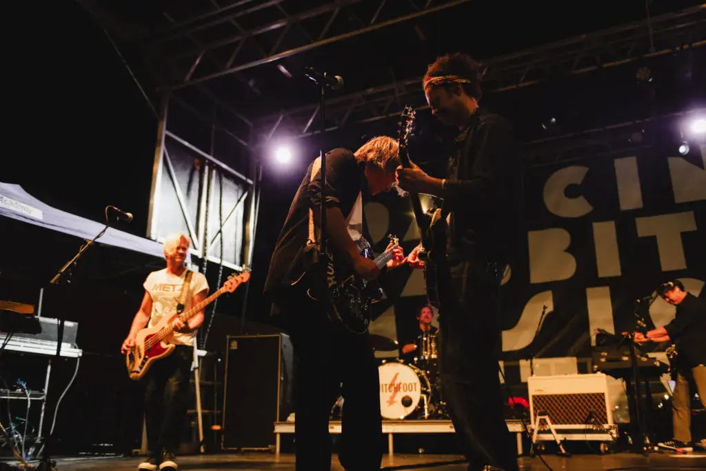 A band performs on stage at night with multiple musicians playing instruments under bright stage lighting.