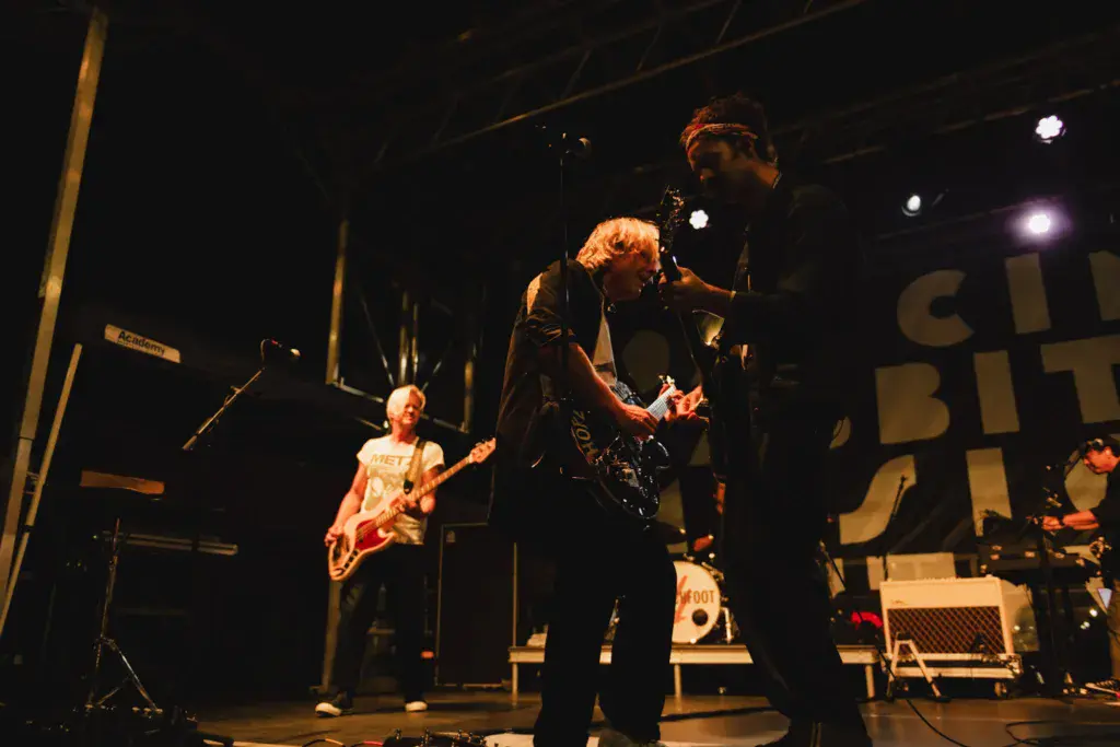 A band performs on stage under warm stage lighting at an indoor venue during the Dancing Rabbit Music Festival.