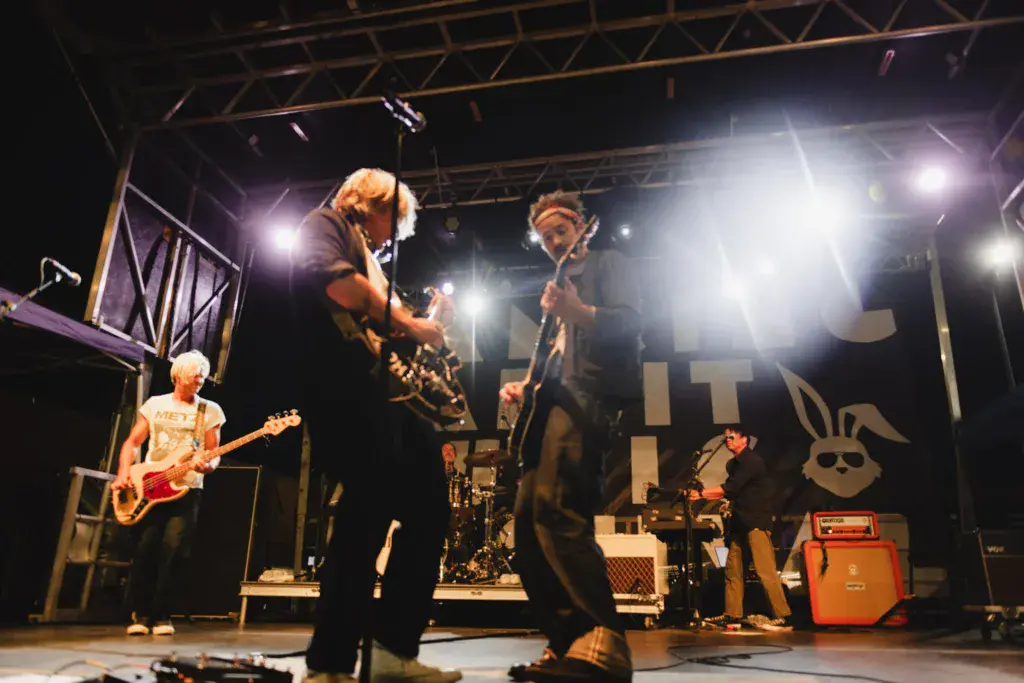 A band performs on a lit stage at night with musicians playing guitars, bass, and drums.