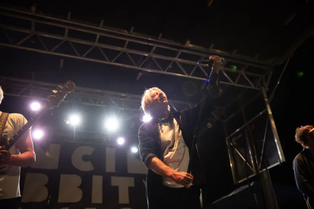 A performer on stage at night under bright stage lights, singing into a microphone with one arm raised.