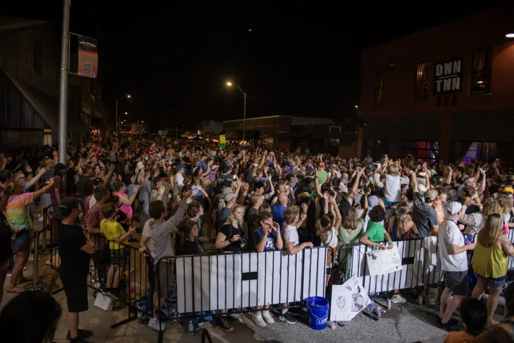 A large crowd of festival attendees gathered in an outdoor urban venue at night, many with hands raised.